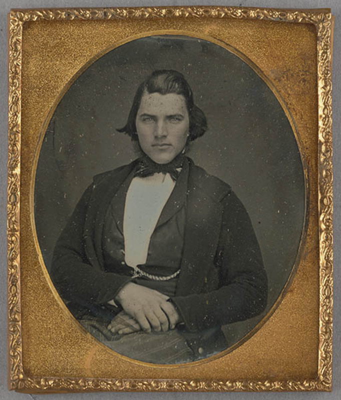 Retrato de medio cuerpo de un hombre joven con mirada directa y ojos claros. Viste indumentaria formal del siglo XIX, compuesta por una chaqueta oscura, camisa blanca de cuello alto y un lazo oscuro anudado al cuello. Se observa una leontina cruzando su chaleco y sus manos reposan tranquilas sobre su regazo, mostrando una pose solemne típica de la época.