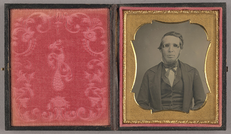 Retrato de estudio de medio cuerpo de un hombre adulto con patillas pronunciadas, vistiendo ropa formal de mediados del siglo XIX que incluye saco oscuro, chaleco y corbata. Destaca el uso de anteojos con cristales oscuros. La imagen está resguardada en un estuche abierto con una cubierta interior de terciopelo rojo decorada con un motivo de pavo real.
