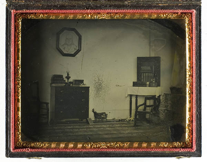 Vista interior de una habitación doméstica de mediados del siglo XIX. La escena incluye una cómoda con libros y una lámpara, un retrato octogonal colgado en la pared y una mesa con mantel blanco. En el suelo se aprecia un juguete pequeño, y a la derecha aparece una figura humana borrosa debido al movimiento durante la exposición prolongada.
