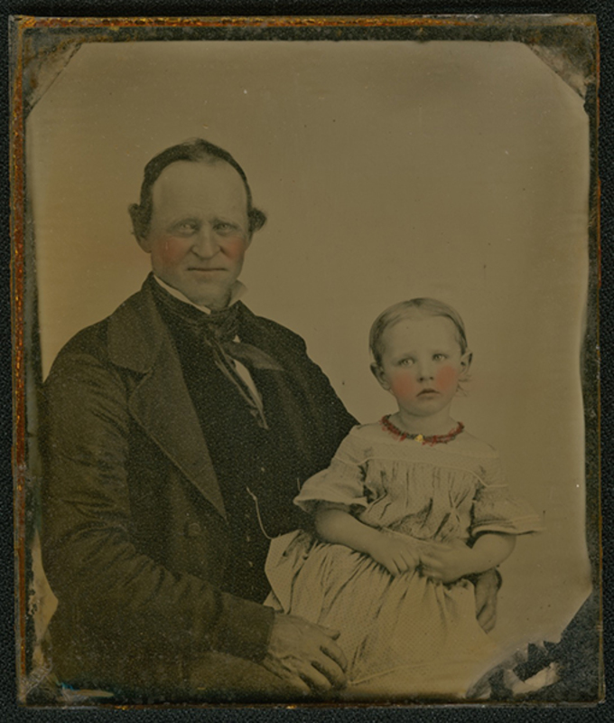 Retrato de estudio de mediados del siglo XIX que muestra a un hombre de mediana edad junto a un infante. El adulto viste prendas formales oscuras con un pañuelo al cuello, mientras el niño porta un vestido estampado y un collar. La imagen destaca por el sutil coloreado manual en las mejillas de ambos sujetos, buscando un efecto realista.