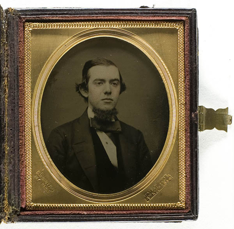 Retrato de estudio de un joven de mediados del siglo XIX, representado de medio cuerpo con una pose formal. El sujeto luce un peinado con raya lateral y una barba de collar (chin curtain). Viste una indumentaria de época que consiste en una levita oscura, chaleco a juego, camisa de cuello alto y un corbatín ancho.