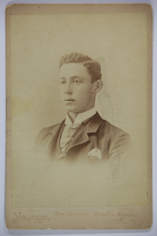 Retrato de estudio de un hombre joven capturado en tres cuartos de perfil. El sujeto viste un atuendo formal compuesto por un saco oscuro con pañuelo de bolsillo, camisa de cuello alto rígido y una corbata ancha de estilo victoriano. La fotografía muestra un acabado en viñeta que difumina los bordes inferiores del retrato sobre un fondo neutro.