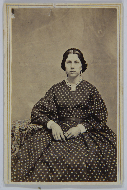 Retrato de estudio de una mujer joven en posición sedente, vestida con un traje de época de color oscuro con patrón de puntos blancos. Luce un collar de perlas con un camafeo central, mangas abullonadas y un tocado de encaje oscuro. La composición es formal, con un fondo neutro y una mesa lateral con mantel ornamental.