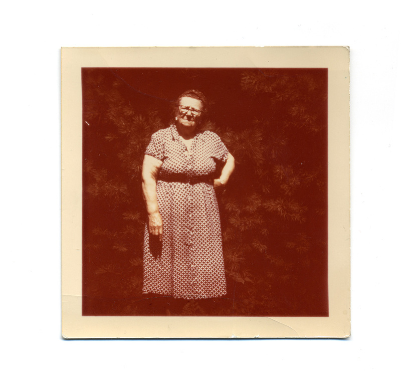 Retrato de una mujer de edad avanzada posando de pie en un entorno exterior frente a un arbusto denso. Viste un vestido estampado de manga corta con botones al frente y anteojos de montura oscura. La composición es un retrato vernáculo típico de la fotografía familiar mexicana de mediados del siglo XX.