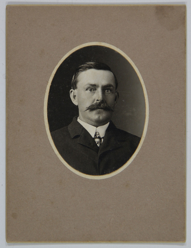 Retrato de estudio de un hombre de mediana edad con un bigote espeso y bien definido. Viste ropa formal de principios del siglo XX, que incluye un saco oscuro, camisa blanca de cuello alto rígido y una corbata con patrón gráfico. La fotografía está contenida en un marco ovalado impreso, montada sobre un soporte de cartón grueso de tono neutro que presenta un sello seco de estudio en la esquina inferior derecha.