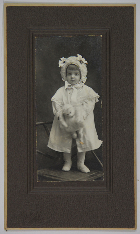 Retrato de estudio de una niña pequeña en posición frontal, ataviada con ropa de invierno que incluye un abrigo claro, un gorro ornamentado con lazos y calzado de lana. La infante sostiene lo que parece ser una muñeca o juguete de felpa. La imagen está capturada en un entorno controlado con iluminación dirigida y un fondo oscuro liso.