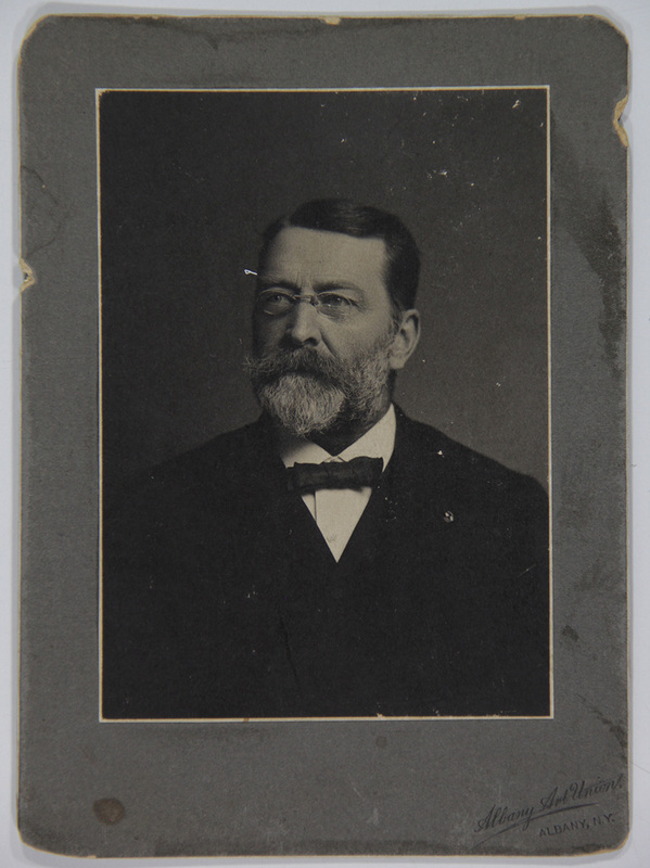 Retrato de estudio de un hombre de edad madura con anteojos redondos, barba densa y bigote entrecano. Viste indumentaria formal compuesta por una chaqueta oscura, camisa blanca de cuello rígido y una corbata de lazo (bow tie) oscura. El sujeto posa de tres cuartos mirando hacia un punto fuera de cámara.