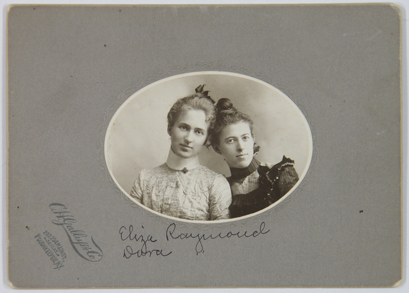 Retrato de estudio de busto de dos mujeres jóvenes. Ambas visten indumentaria formal de la era victoriana tardía, con blusas de cuello alto y detalles ornamentales en encaje y seda. La imagen está centrada en un formato ovalado sobre un soporte de cartón que incluye el sello del fotógrafo y una identificación manuscrita de los sujetos.