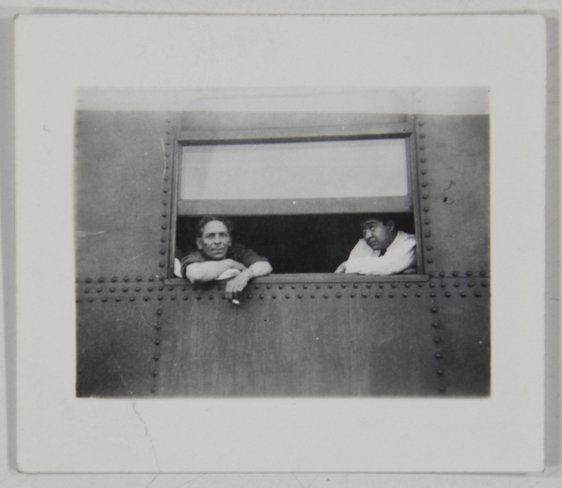 Retrato de dos hombres asomados por la ventana de un vagón ferroviario construido con placas de metal remachadas. El hombre de la izquierda viste una camisa oscura y observa directamente a la lente con los brazos apoyados en el marco, mientras que el hombre a su derecha, de complexión más robusta y camisa clara, mantiene la mirada baja en una actitud reflexiva durante lo que parece ser una parada del trayecto.
