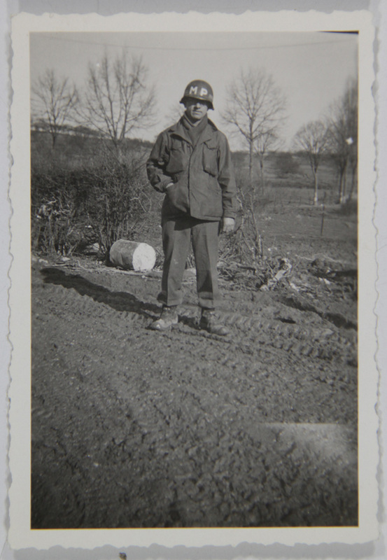 Retrato de cuerpo completo de un hombre joven uniformado como Policía Militar (MP). El sujeto posa de pie frente a un paisaje rural invernal con árboles desnudos y un suelo lodoso que muestra huellas de neumáticos de gran tamaño. Viste una chaqueta de campo M-1943, pantalones de lana y casco reglamentario marcado, situando la escena probablemente en Europa a mediados de la década de 1940.