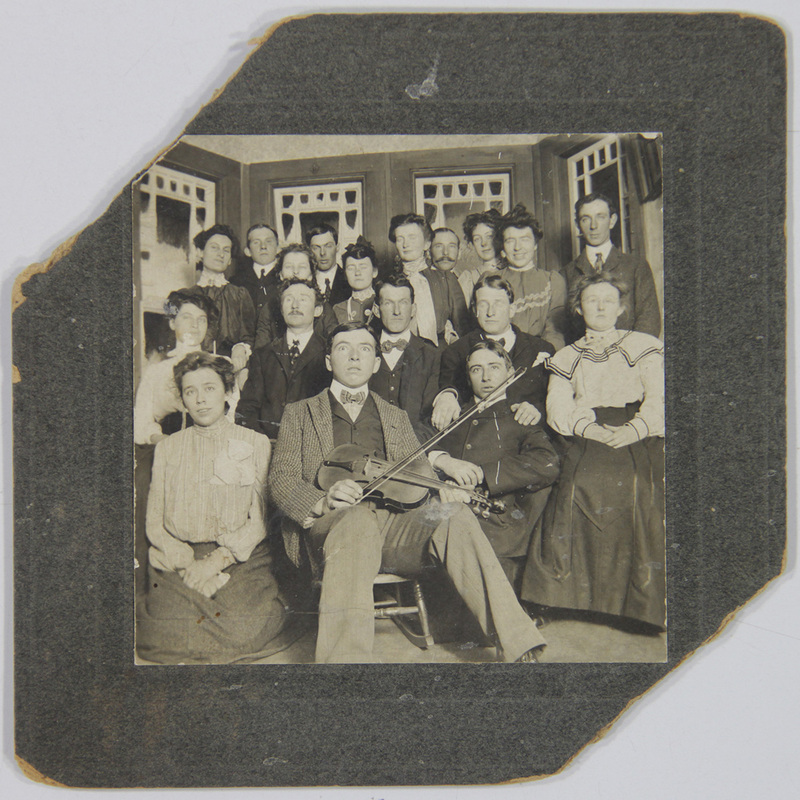 Un retrato grupal de quince individuos, hombres y mujeres, posando de manera formal en un entorno interior decorado con paneles acristalados. El foco central es un hombre sentado que sostiene un violín. Los integrantes presentan una vestimenta típica de la clase media o alta de principios del siglo XX, sugiriendo un evento social o familiar.