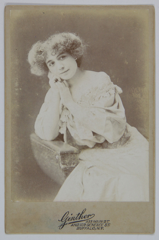 Retrato de estudio en plano medio de una mujer joven con cabello corto y rizado. La figura aparece apoyada sobre un mueble de utilería, sosteniendo su rostro con ambas manos en una pose introspectiva. Viste un vestido de tono claro con un prominente cuello de encaje, típico de la estética de finales de la época victoriana.