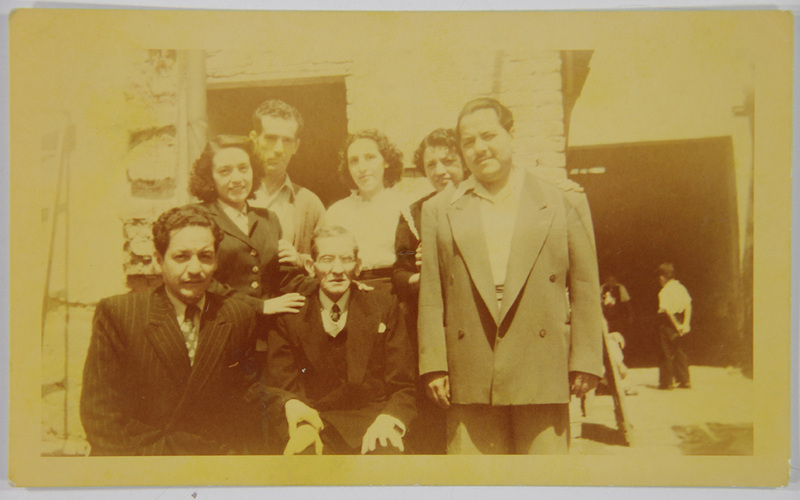 Retrato grupal de siete personas, cuatro hombres y tres mujeres, posando de manera formal en lo que parece ser un patio o la entrada de una edificación con muros de piedra y ladrillo. Los sujetos visten ropa de corte clásico de mediados del siglo XX; destaca un hombre de edad avanzada sentado en el centro y una figura infantil visible al fondo a la derecha. La escena evoca una reunión familiar o social en un entorno doméstico o de trabajo artesanal.