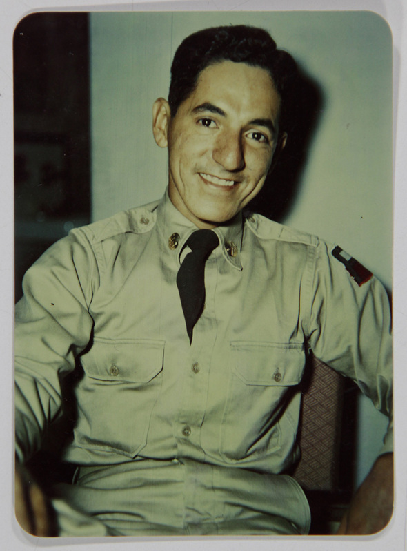 Retrato de estudio de un hombre joven vestido con uniforme militar estadounidense de color caqui y corbata negra. En su manga izquierda porta un parche de una división acorazada. El sujeto posa sentado con una expresión amable, representando la iconografía clásica de los soldados de mediados del siglo XX, posiblemente de origen mexicano-estadounidense.