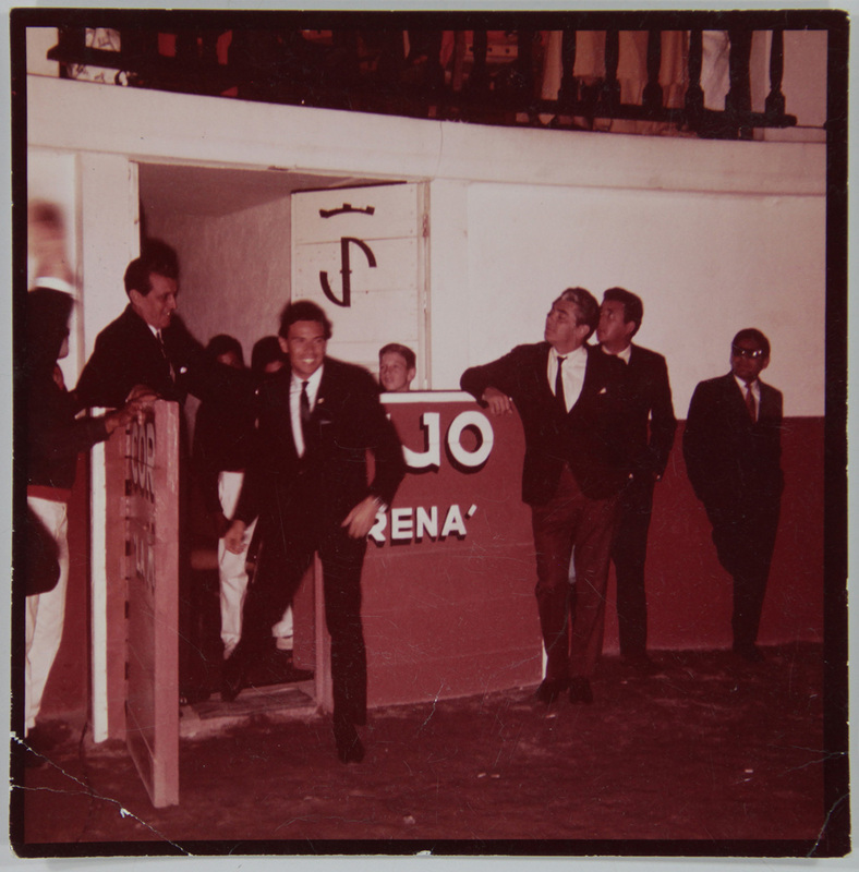 Escena capturada en el callejón de una plaza de toros en México, probablemente en los años 60. Se observa a varios hombres con trajes formales y corbatas delgadas; uno de ellos camina hacia el ruedo mientras otros observan apoyados en un burladero donde se lee parcialmente la palabra 'ARENA'. Al fondo destaca el hierro de una famosa ganadería sobre una puerta.