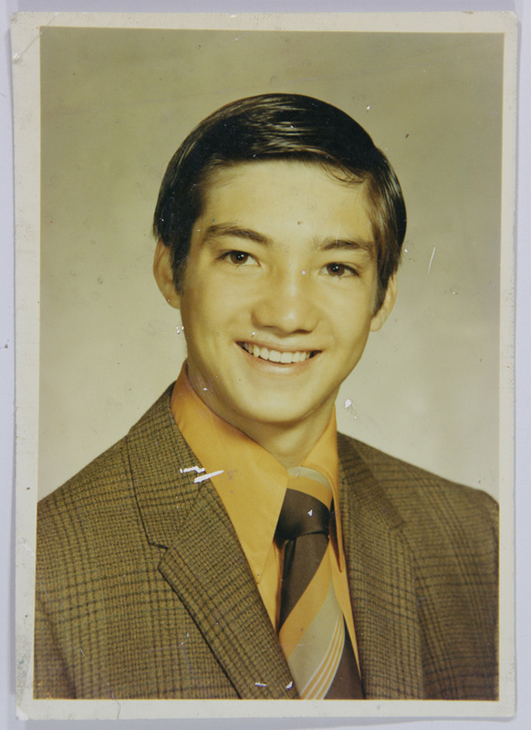 Retrato de estudio individual de un hombre joven en encuadre de busto. El sujeto posee rasgos mestizos, cabello oscuro peinado hacia un lado y expresión sonriente. Viste un atuendo formal compuesto por un saco con diseño de cuadros pequeños, camisa naranja brillante de cuello largo y corbata ancha con franjas diagonales en tonos tierra, típica de la moda urbana mexicana de los años 70.