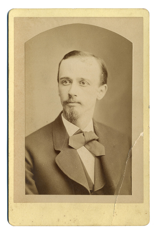 Retrato de estudio en plano medio de un hombre joven con mirada dirigida hacia su derecha. El sujeto viste un traje formal oscuro de solapas anchas, camisa de cuello rígido y un corbatín de lazo prominente. Su fisonomía destaca por una frente amplia, bigote delgado y una barba de tipo perilla cuidadosamente recortada.