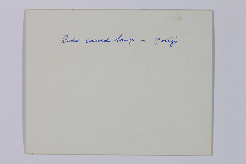 Se trata del reverso de una fotografía histórica con una anotación manuscrita en tinta azul que reza 'Dads' carved lamp ~ Pollys'. El texto indica que la imagen en el anverso documenta un objeto decorativo o artesanal dentro de un contexto doméstico y familiar, probablemente para fines de inventario o recuerdo personal.