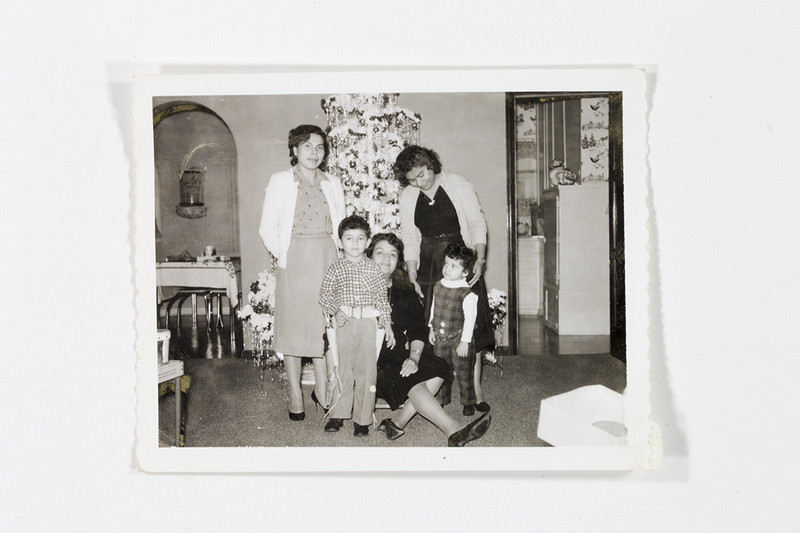 Retrato grupal de una familia en un interior doméstico durante una celebración navideña. Tres mujeres de distintas edades y dos niños pequeños posan frente a un árbol de Navidad decorado con festones y esferas. La escena muestra una estancia con un arco de medio punto y una vista hacia una cocina con papel tapiz decorativo, capturando la estética hogareña mexicana de mediados del siglo XX.