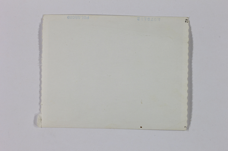 Vista del reverso de una fotografía instantánea que muestra el sello de autenticidad de la marca Polaroid. La superficie de papel presenta una pátina amarillenta por el envejecimiento natural y marcas de producción técnica en los márgenes. Representa el uso de tecnología de revelado inmediato, común en la documentación fotográfica cotidiana y periodística en México durante la segunda mitad del siglo XX.