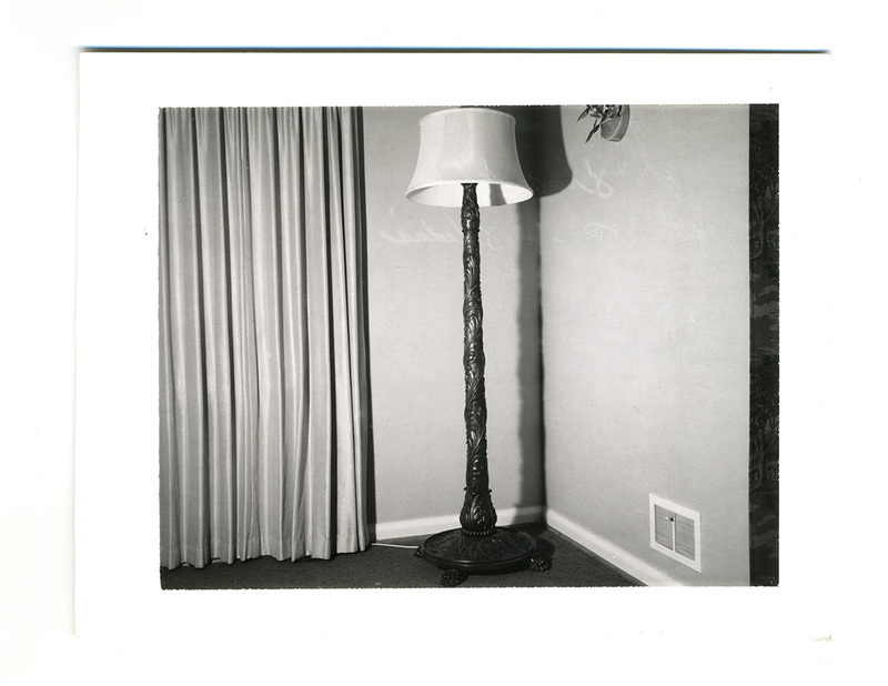 Una fotografía de un rincón interior que destaca una lámpara de pie ornamentada con fuste tallado y una pantalla de tela. A la izquierda se observan cortinas de pliegues verticales y a la derecha una pared con una rejilla de ventilación cerca del suelo, reflejando el diseño decorativo de mediados del siglo XX.