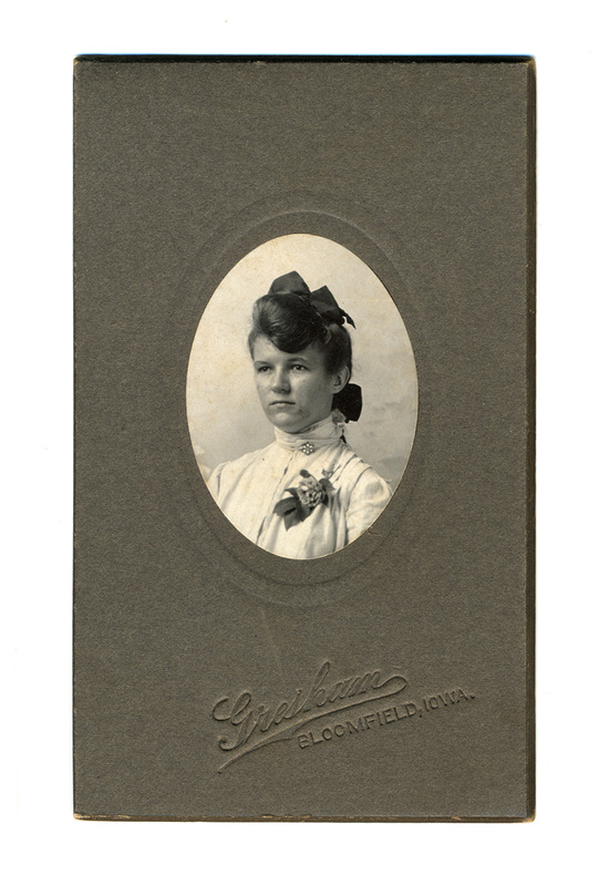 Retrato de estudio de una joven mujer en plano medio corto, presentado en un óvalo. Viste una blusa clara de cuello alto con encajes, un broche central y un ramillete floral. Su cabello está recogido con un lazo oscuro de gran tamaño, característico de la estética de principios del siglo XX.