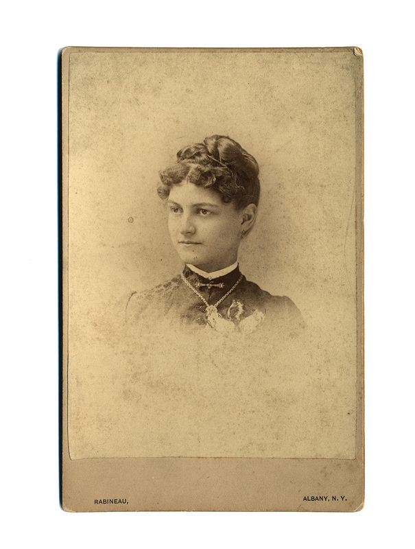 Retrato de estudio en plano medio corto de una mujer joven. La figura posa de tres cuartos con la mirada dirigida hacia un lateral. Viste una prenda oscura de cuello alto, decorada con un collar metálico y un broche. Su cabello está recogido en un moño elaborado con flequillo de rizos cortos sobre la frente, siguiendo la moda victoriana tardía de la década de 1880.