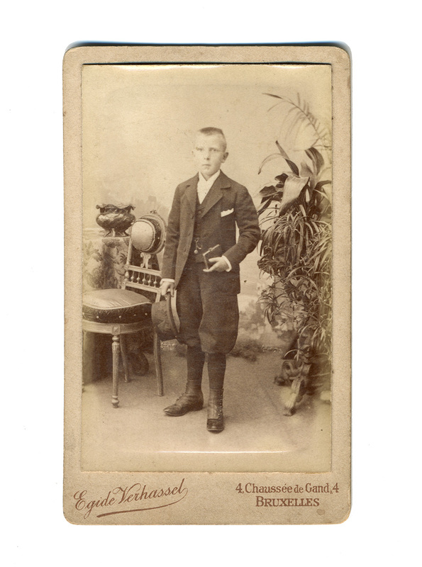 Retrato de estudio de un niño de pie, vestido con un traje formal oscuro de tres piezas: chaqueta, chaleco y pantalones tipo bombacho. El sujeto sostiene un sombrero en su mano derecha y un pequeño objeto rectangular, posiblemente un libro, en la izquierda. El entorno incluye una silla decorativa, una mesa con mantel floral y plantas ornamentales de gran tamaño al fondo.