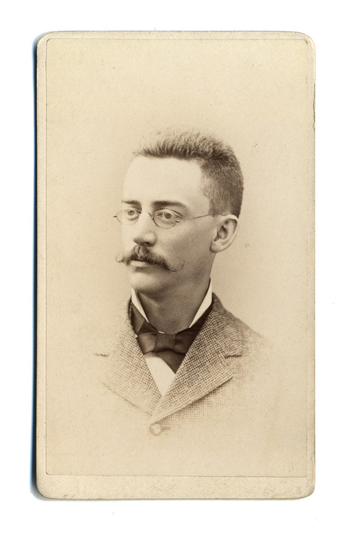 Retrato de estudio de un hombre joven captado de tres cuartos. El sujeto posee cabello corto, un bigote denso y utiliza anteojos circulares. Viste una chaqueta de textura gruesa con solapas pequeñas, una camisa de cuello alto y un corbatín oscuro. La composición y el fondo neutro son típicos de la retratística comercial decimonónica.