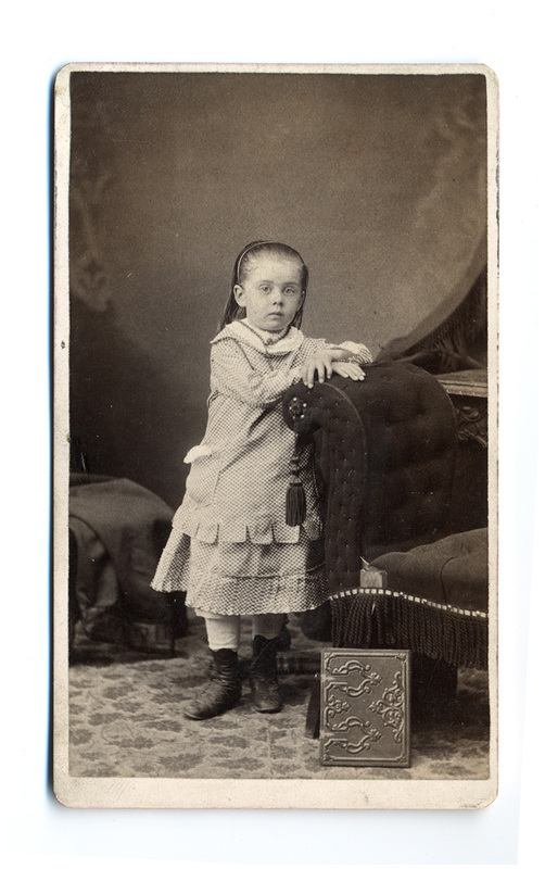 Retrato de estudio de una infante de pie, ataviada con un vestido de patrón cuadrangular con volantes y botas altas de piel. Se apoya en una silla tapizada de estilo victoriano con flecos decorativos. La escena, capturada ante un telón pintado, incluye un álbum ornamental en el suelo, reflejando las convenciones estéticas y sociales del retrato burgués de finales del siglo XIX.