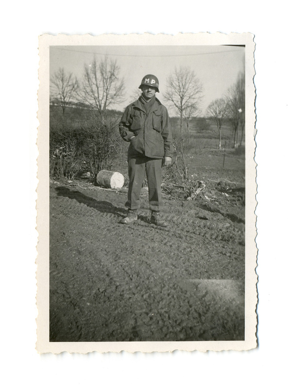 Retrato de cuerpo entero de un soldado de la Policía Militar estadounidense en un entorno rural lodoso. El individuo viste una chaqueta de campo M-1943, casco con las siglas 'MP' y botas de combate. La escena, capturada en exteriores con árboles caducifolios al fondo, evoca el periodo de la Segunda Guerra Mundial o la posguerra inmediata.