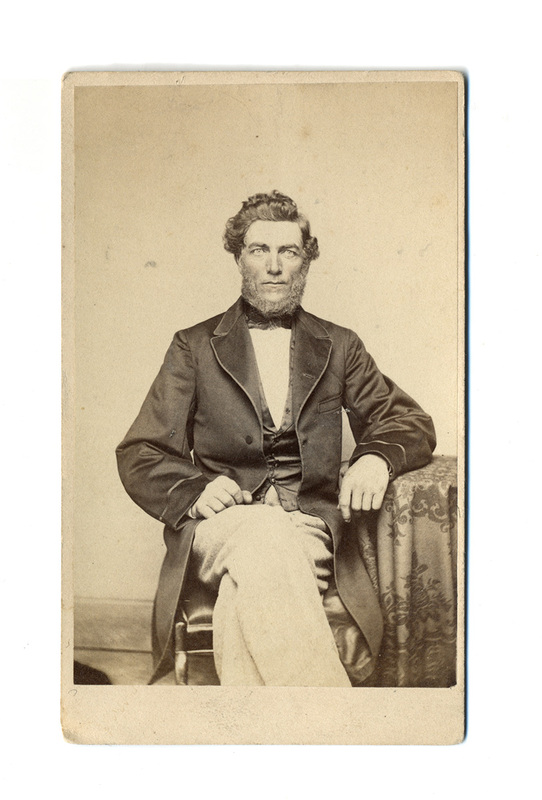 Retrato de estudio de un hombre adulto con cabello ondulado y barba abundante sin bigote. Viste una indumentaria formal compuesta por una levita oscura, chaleco abotonado, camisa blanca de cuello alto y corbatín oscuro. El sujeto aparece sentado en una pose formal, con el brazo izquierdo apoyado sobre una mesa cubierta con un mantel de motivos decorativos florales.
