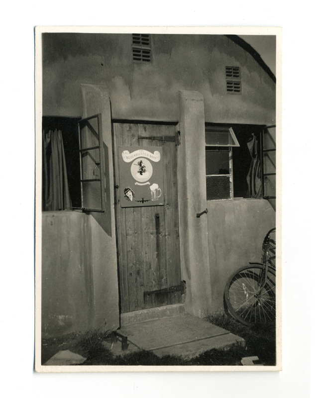 Vista exterior de la entrada a un alojamiento militar, probablemente un cuartel de tripulación aérea durante la Segunda Guerra Mundial. En la puerta de madera destaca un cartel titulado 'The Blockbuster Boys', ilustrado con arte gráfico que incluye el rostro de una mujer, un bombardero y una cerveza. Una bicicleta se encuentra apoyada contra la pared lateral.