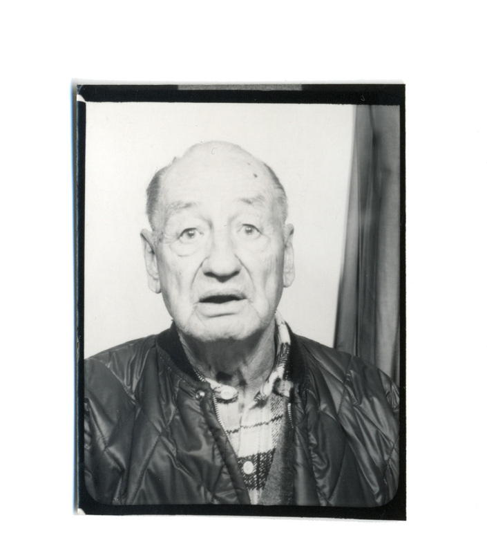 Retrato en plano medio de un hombre de edad avanzada capturado en un formato de identificacion. El sujeto muestra una expresion de sorpresa con la boca ligeramente abierta, vistiendo una chaqueta acolchada sobre una camisa de cuadros. El fondo es neutro con el detalle de una cortina a la derecha, indicando un entorno de cabina fotografica o estudio basico.