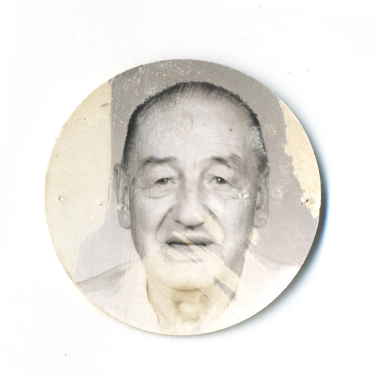 Retrato de busto de un hombre adulto mayor, capturado de frente con una expresión neutra. El sujeto presenta rasgos de envejecimiento marcados y viste una camisa de tono claro. La imagen está recortada en forma circular y muestra daños por desprendimiento de emulsión y marcas de grapas o tachuelas en los extremos laterales.