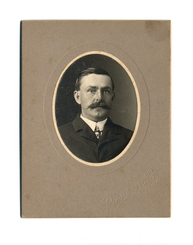 Retrato de estudio de un hombre adulto capturado a principios del siglo XX. El sujeto luce un bigote tupido tipo manubrio, viste traje oscuro de solapa ancha, camisa blanca con cuello rígido y corbata con un alfiler ornamental. La composición es un busto frontal dentro de un marco ovalado sobre un soporte rígido.