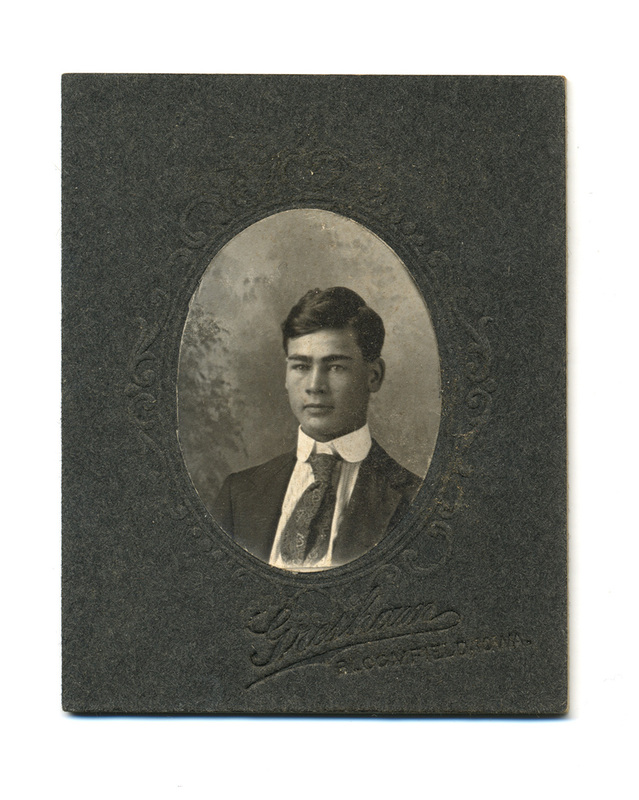 Retrato de estudio de un hombre joven de cabello oscuro y rasgos marcados, capturado en una pose de tres cuartos. Viste un traje formal con una camisa de cuello rígido y una corbata de seda con intrincados motivos ornamentales. La fotografía está contenida en un óvalo impreso sobre una montura de cartón grisáceo decorada con filigranas en relieve, característica de los estudios fotográficos comerciales estadounidenses de la época.