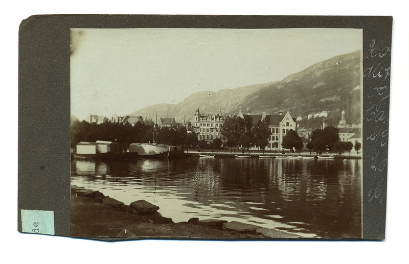 Vista panorámica de un lago urbano rodeado de edificaciones de estilo europeo a finales del siglo XIX. Se observan pequeñas embarcaciones blancas alineadas en la orilla y botes de mayor calado a la izquierda. El paisaje está dominado por una montaña escarpada al fondo bajo una iluminación natural diurna.