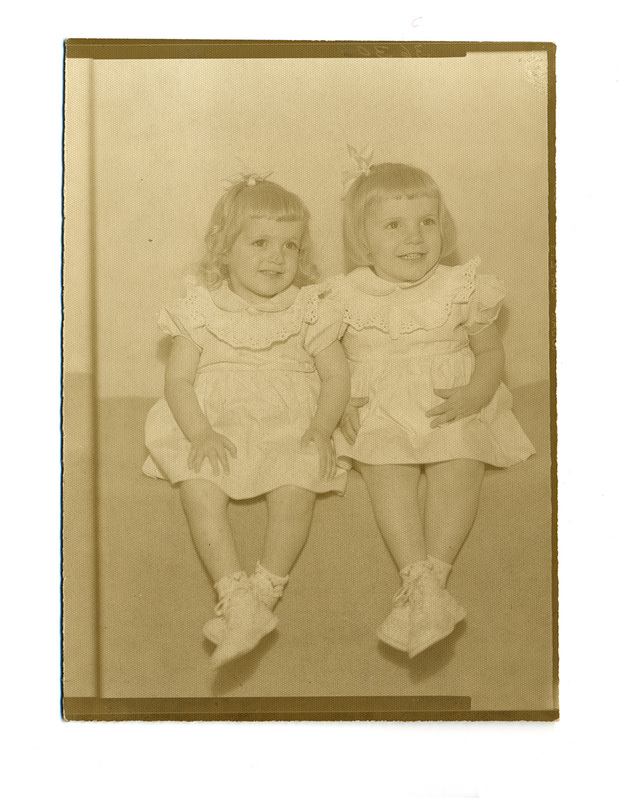 Retrato de estudio en plano medio de dos niñas pequeñas sentadas sobre un pretil o superficie elevada. Ambas lucen peinados con pequeños moños laterales y visten atuendos idénticos compuestos por vestidos claros con detalles de encaje en el cuello y mangas, calcetas blancas y zapatos de horma clásica. La iluminación es frontal y suave, propia de la fotografía de gabinete familiar de mediados del siglo XX.