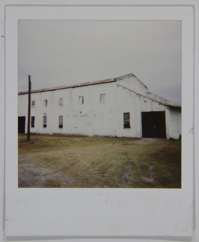 Vista exterior de un edificio alargado de mampostería blanca, posiblemente una bodega o instalación industrial rural en México. La estructura presenta una techumbre a dos aguas, múltiples ventanas pequeñas alineadas y un acceso principal oscuro en el extremo derecho. El entorno se compone de un terreno llano con vegetación escasa bajo un cielo nublado.