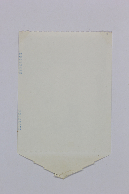 Vista posterior de una fotografía instantánea Polaroid. Se observa un soporte de papel color crema con la marca del fabricante y un número de serie estampados lateralmente en tinta azul. El diseño incluye bordes troquelados y una pestaña en forma de flecha en el extremo inferior, elementos funcionales del antiguo proceso de revelado por difusión-transferencia manual.