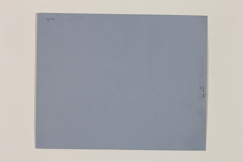 La imagen muestra el reverso de una película instantánea de formato profesional. No presenta una escena fotográfica visible, sino una superficie plana y uniforme de color gris azulado. En el borde derecho se observa un sello técnico de fábrica con el modelo del producto, y en la esquina superior izquierda se aprecia un número '1' escrito manualmente.