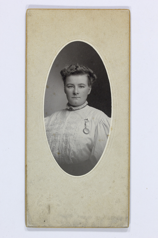 Retrato de estudio de una mujer joven capturado en un formato vertical. La figura aparece de medio cuerpo, luciendo una blusa blanca con detalles de bordado y un broche circular prendido al pecho. Su cabello está peinado con ondas hacia atrás, y mantiene una expresión formal frente a un fondo neutro degradado.