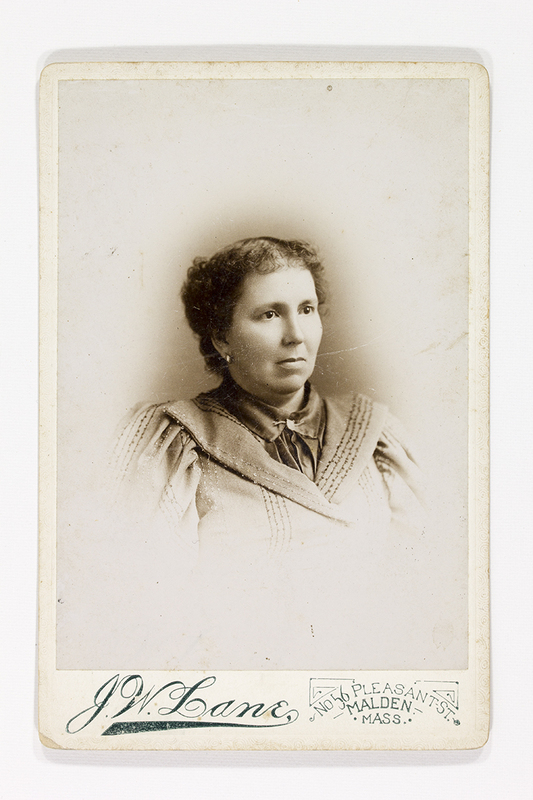 Retrato de estudio de una mujer adulta de tez clara con el cabello recogido. Viste una blusa de cuello alto con aplicaciones decorativas lineales y hombros ligeramente estructurados, moda típica de finales del siglo XIX. La composición utiliza un efecto de viñeta que difumina el cuerpo hacia la base del soporte, centrando la atención en el rostro de la retratada.