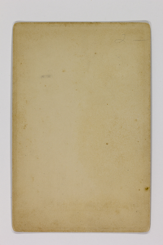 Esta imagen muestra el reverso o verso de una fotografía histórica. Se aprecia un soporte de cartón rígido con pátina de envejecimiento, esquinas redondeadas y diversas manchas de humedad y oxidación (foxing). En la esquina superior derecha se observa una breve anotación manuscrita a lápiz con el número '2'.
