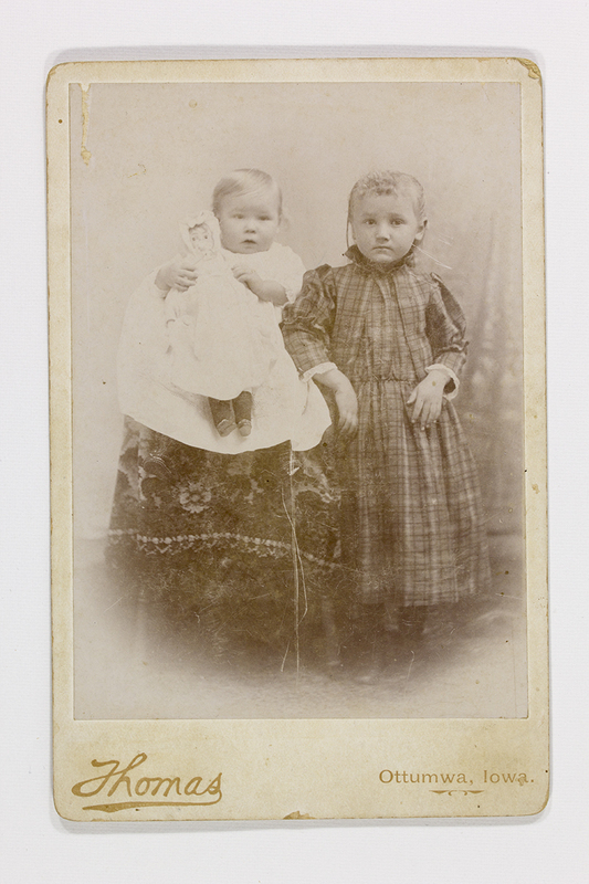 Retrato de estudio de dos niños capturado en Ottumwa, Iowa. A la izquierda, un bebé sentado sobre un regazo cubierto por una tela estampada sostiene una muñeca; a su lado, una niña de pie viste un vestido de tela escocesa con cuello fruncido. Ambos presentan una expresión seria, típica de la fotografía de estudio de finales del siglo XIX, diseñada para conmemorar la infancia.