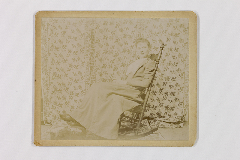 Retrato de estudio de una mujer joven sentada en una mecedora de madera torneada. La sujeto viste un atuendo formal de finales del siglo XIX, compuesto por una falda larga y un saco entallado. La escena está dispuesta frente a un telón con motivos florales, sobre un tapete decorativo y un pequeño descansapiés.