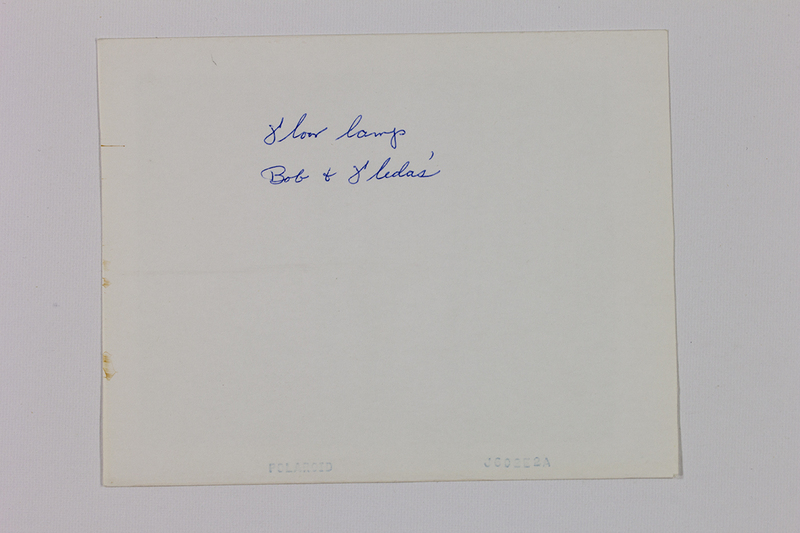 Reverso de una fotografía instantánea con una anotación manuscrita en tinta azul que reza 'floor lamp, Bob & Leda's'. El documento presenta el sello de fábrica 'POLAROID' y un código de producción 'J602E2A' en el borde inferior, además de pequeñas manchas de oxidación o residuos de adhesivo en el margen izquierdo.