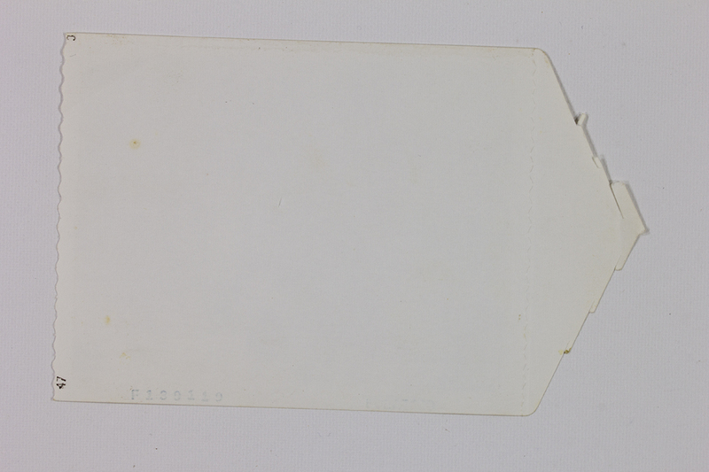 Reverso de una impresión fotográfica de mediados del siglo XX que muestra un borde lateral con acabado festoneado y diversas marcas de catalogación de laboratorio. La pieza presenta una forma irregular y signos de deterioro natural como manchas de oxidación y amarillamiento del papel, conservando códigos numéricos impresos que servían para la identificación industrial de la copia.