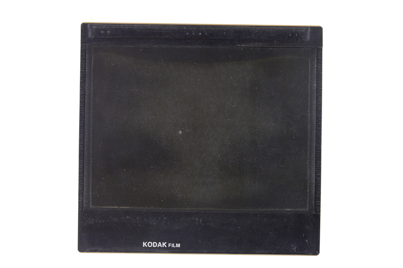 Esta imagen presenta una hoja de película fotográfica de gran formato fabricada por Kodak. Se observa una superficie densa y oscura, carente de detalles figurativos claros, con signos evidentes de deterioro físico como rayaduras y motas de polvo. El borde inferior muestra la marca del fabricante, característica del material fotosensible profesional utilizado en archivos históricos.